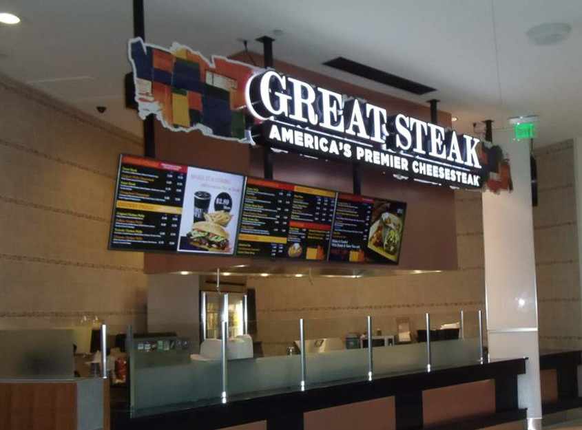 A photograph of the colorful sign of great steak, white neon text letters against a multi-colored collage backdrop